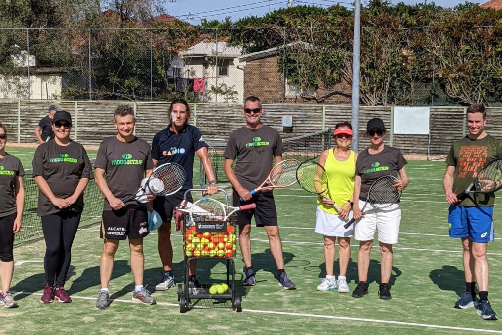 Mood Active trainers and participants on a tennis court, holding rackets and standing by a basket of tennis balls.